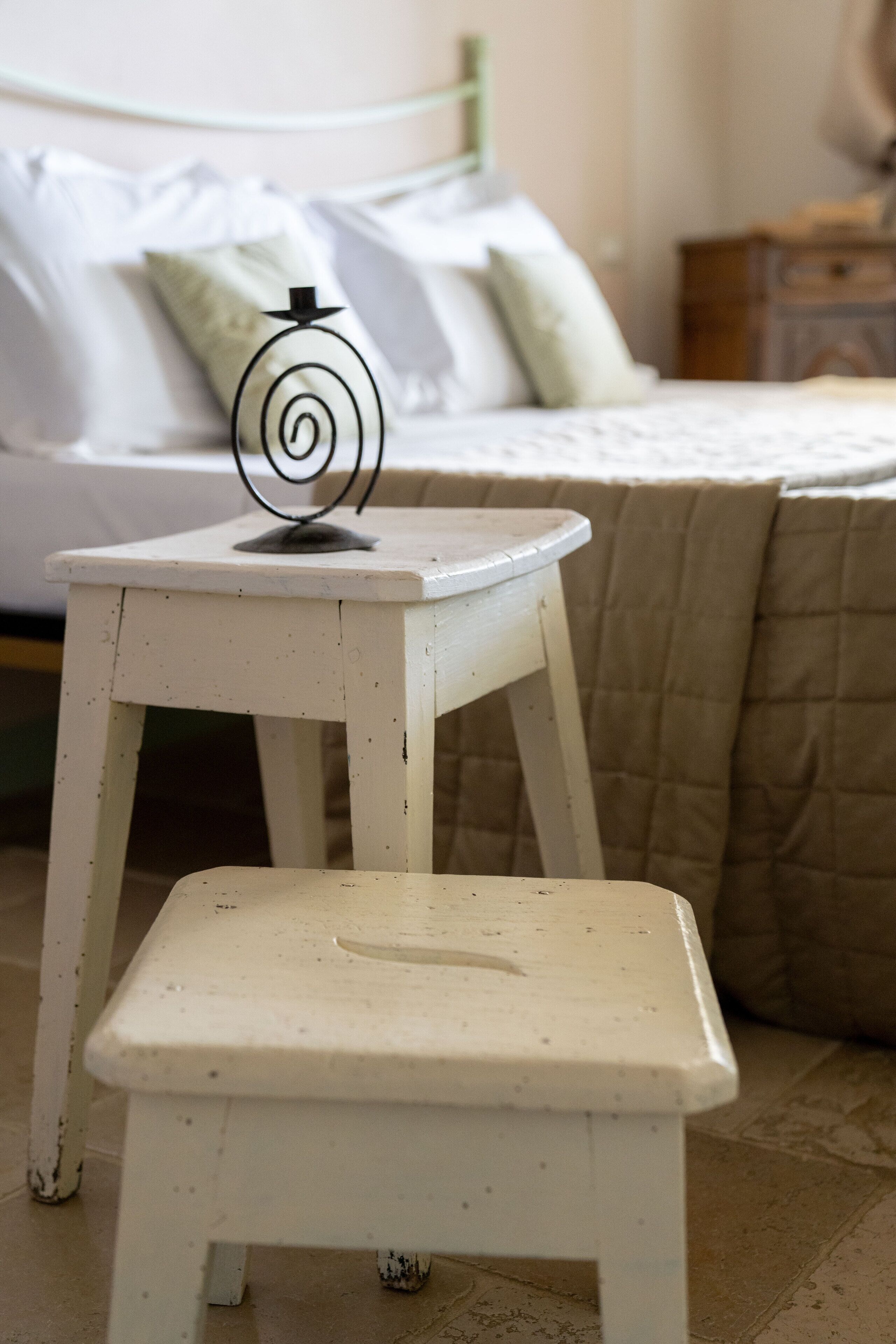 A small white stool with a metal spiral decoration on top, placed in front of a bed with white bedding and a beige quilt.