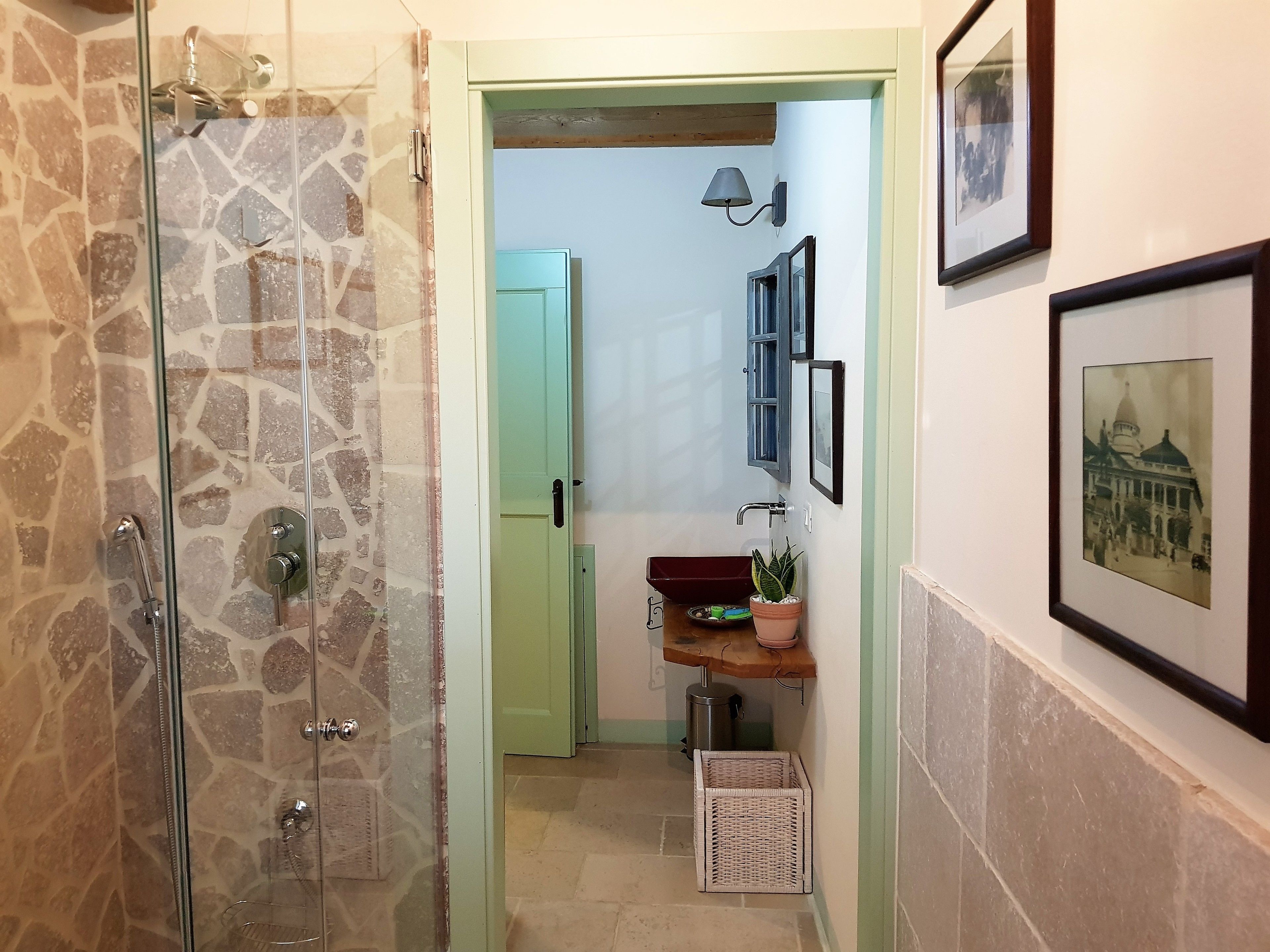 Ein Badezimmer mit Duschkabine aus Glas, einer grünen Tür zu einem Flur, einem Waschbecken mit Spiegel und gerahmten Bildern an der Wand.