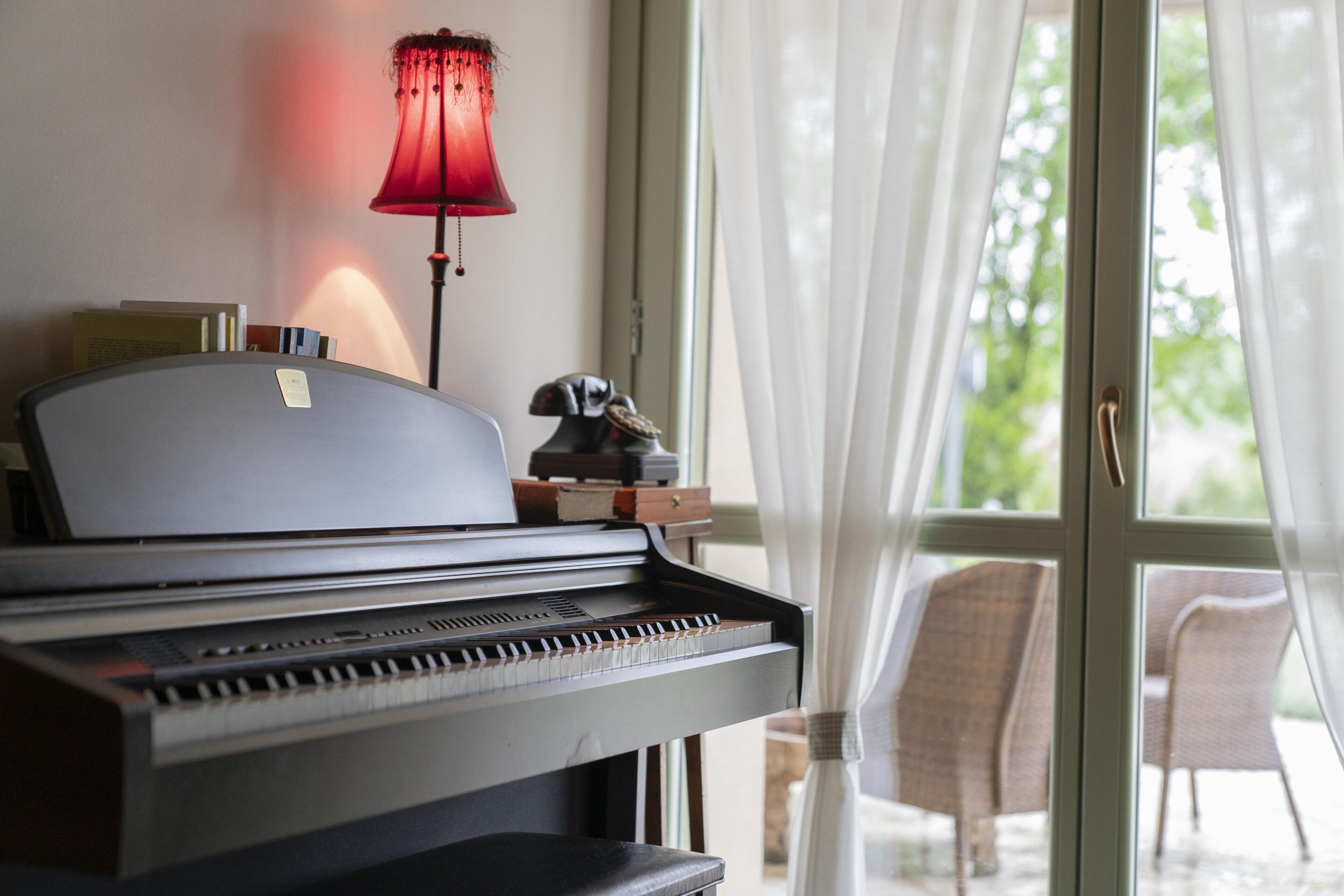 Un piano avec des partitions, une lampe et une vue sur l’extérieur par une fenêtre ornée de rideaux.