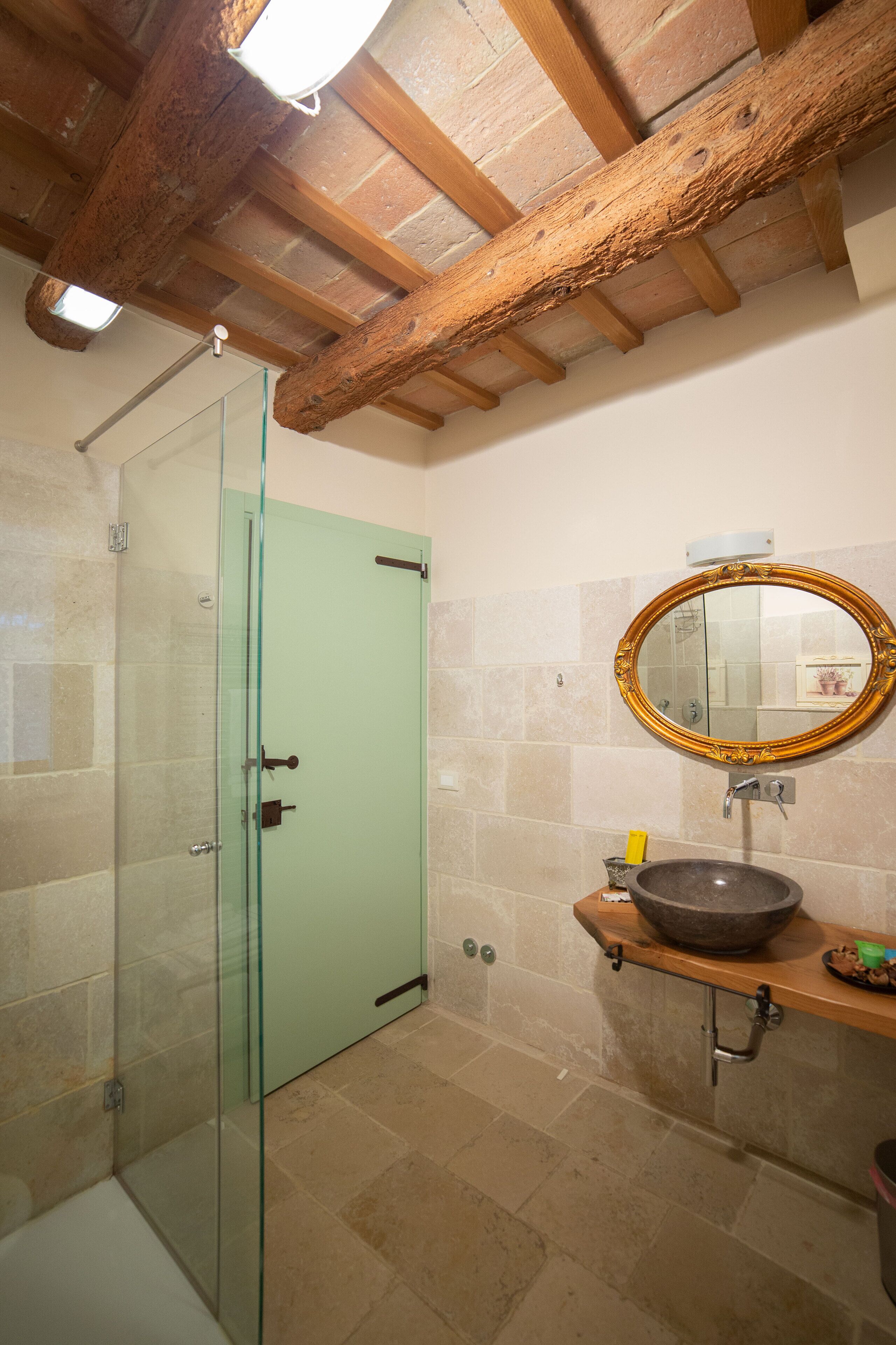 A bathroom with a glass shower enclosure, a green door, a wooden ceiling, a round mirror, and a stone sink.