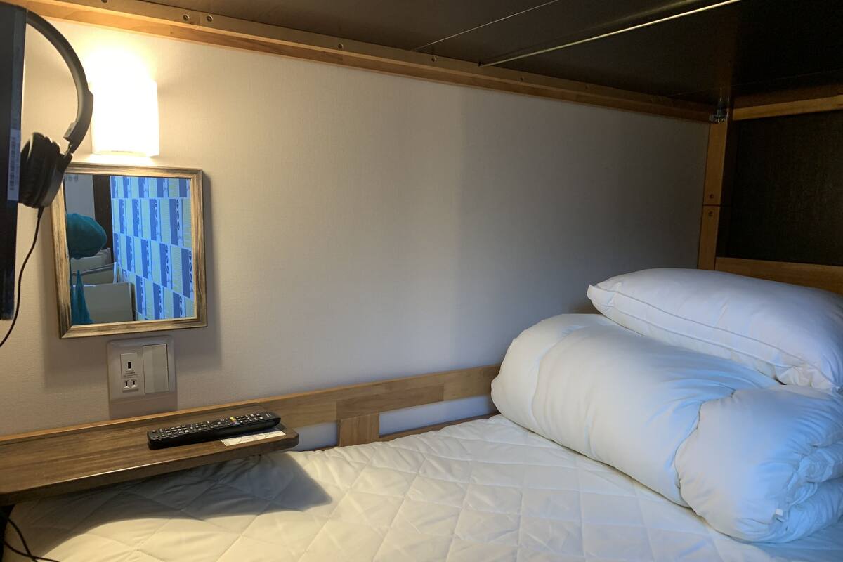 A neatly made bed with white bedding, a bedside table with a remote control, a wall-mounted lamp, and a mirror reflecting part of the room.
