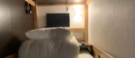 A small, dimly lit room with a bed, a flat-screen TV mounted on the wall, and a wooden headboard.