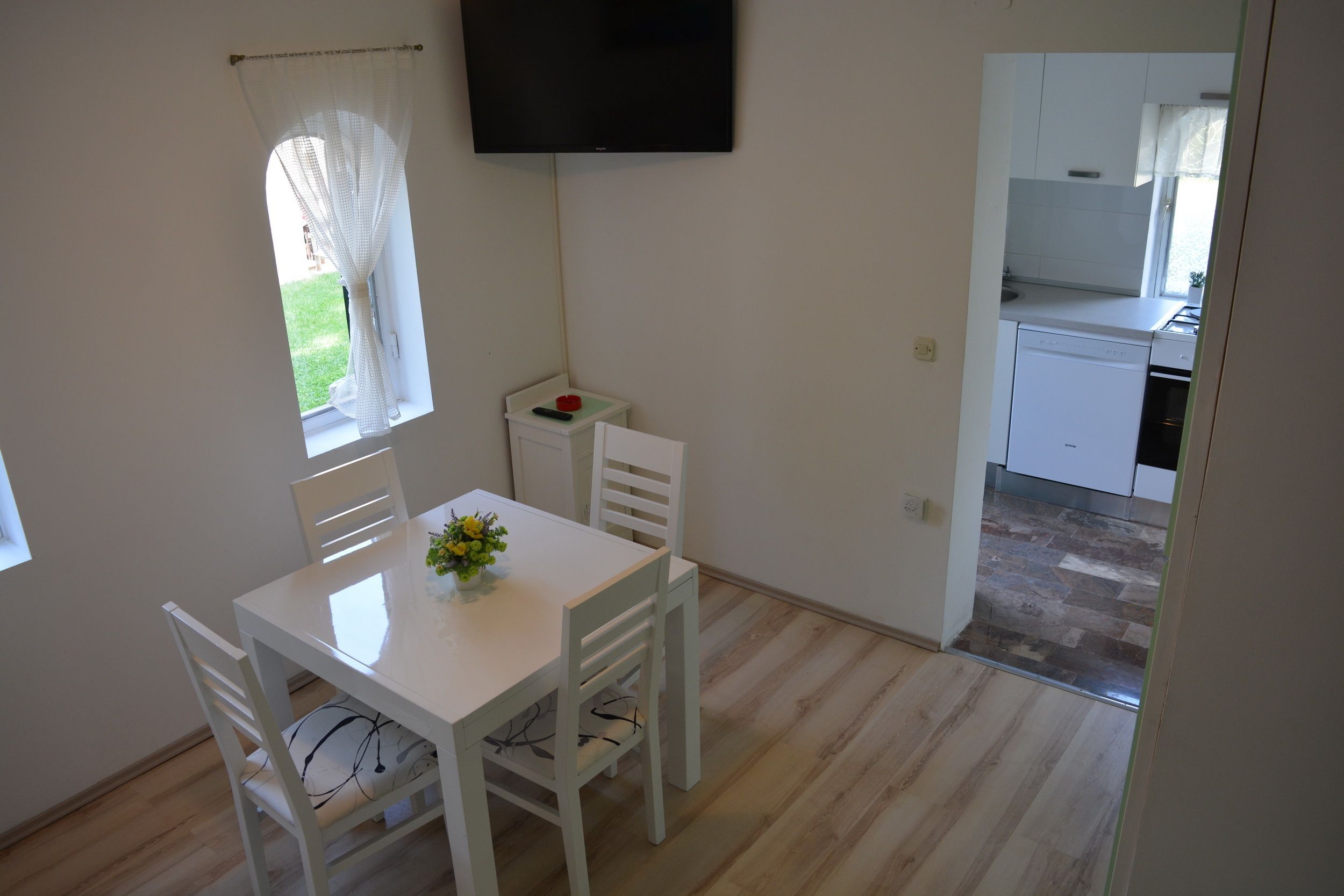 Ein kleiner, sauberer Raum mit Esstisch, Stühlen, einem an der Wand befestigten Fernseher und Blick in eine Küchenzeile.