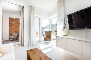 Ein modernes Wohnzimmer mit großem Fernseher, einem Holztisch, einem Korbstuhl und Blick durch eine Glastür nach draußen.