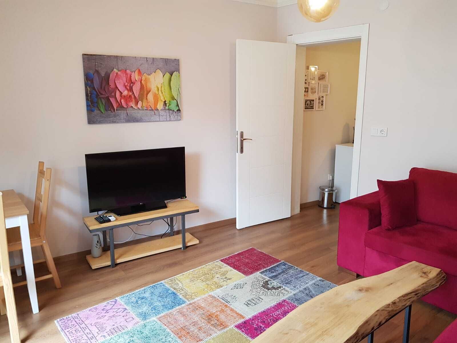 A living room with a flat-screen TV on a wooden stand, a red sofa, a colorful rug, and a wooden bench.