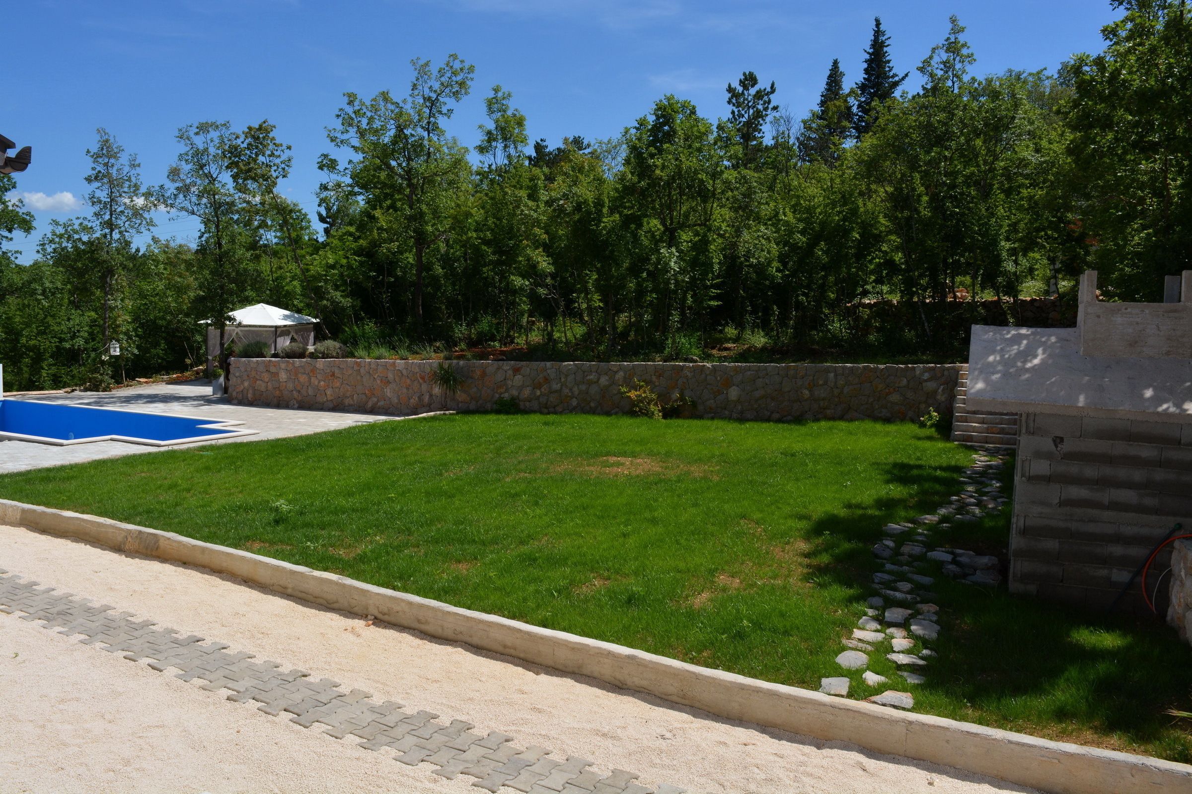 A backyard with a swimming pool, a stone wall, and a paved area.
