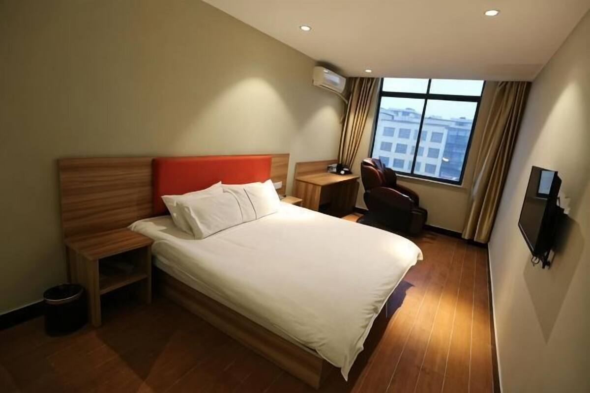 Habitación de hotel con cama, mesita de noche, una silla, televisor y ventana con vistas a edificios.