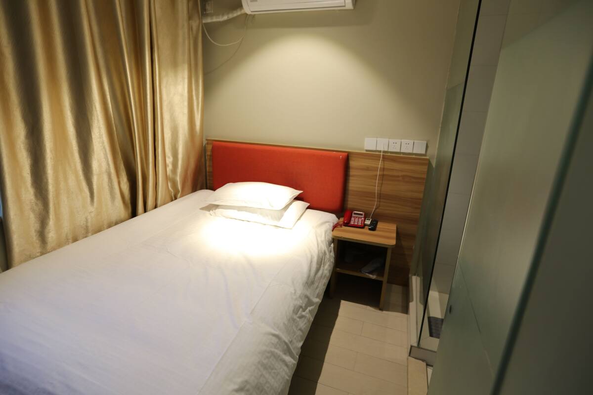 Una cama individual con sábana blanca y cabecera roja, una mesita de noche con teléfono rojo, y un aire acondicionado instalado en la pared.