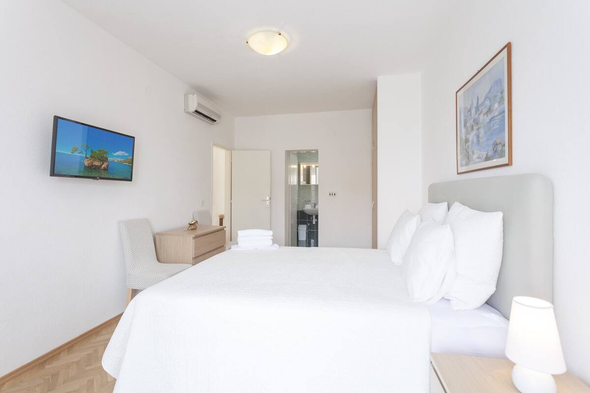 A hotel room with a large bed, a bedside table with a lamp, a chair, a desk, and a painting on the wall.