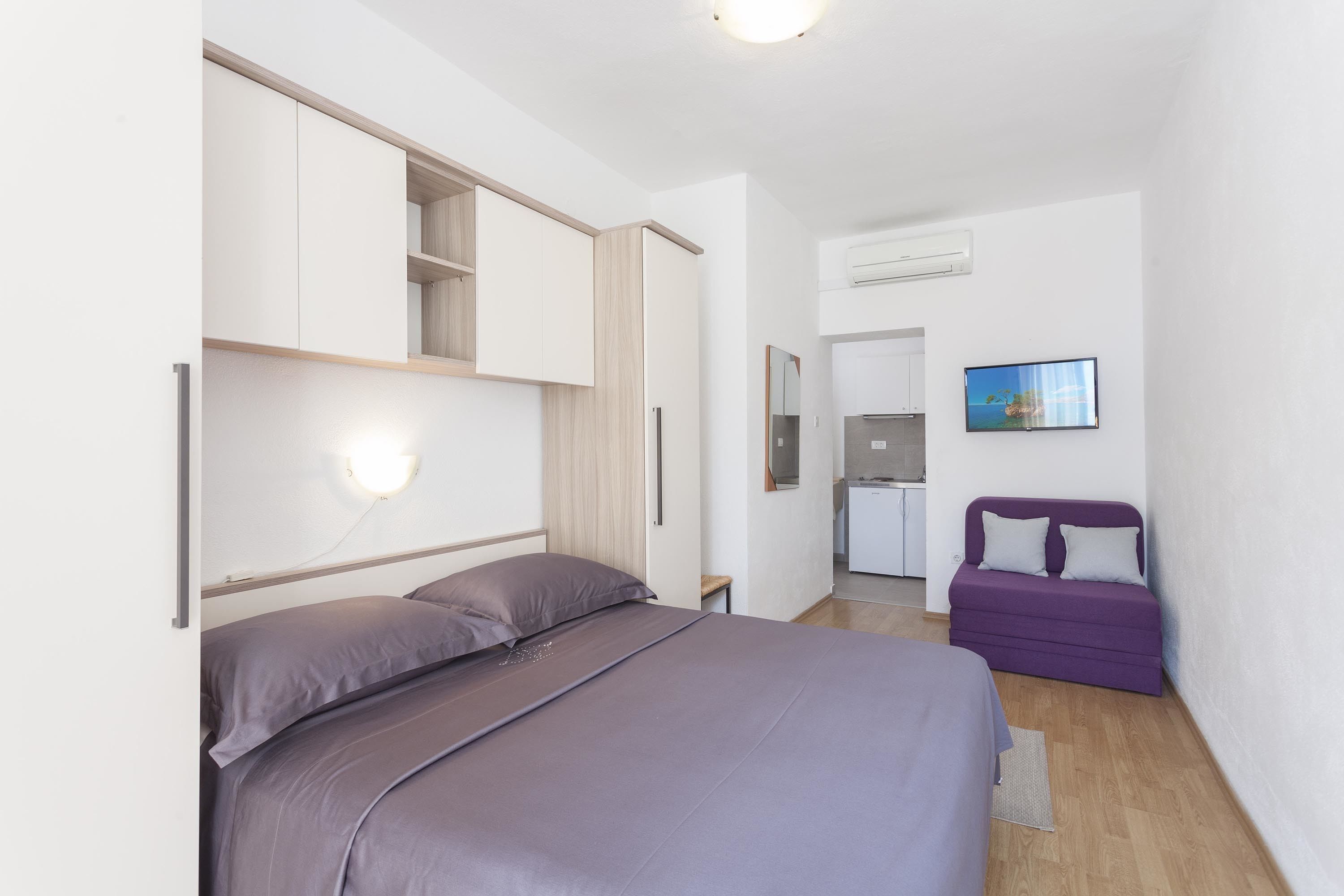 A bedroom with a bed, a built-in wardrobe, a small table, a purple sofa, and a TV on the wall.