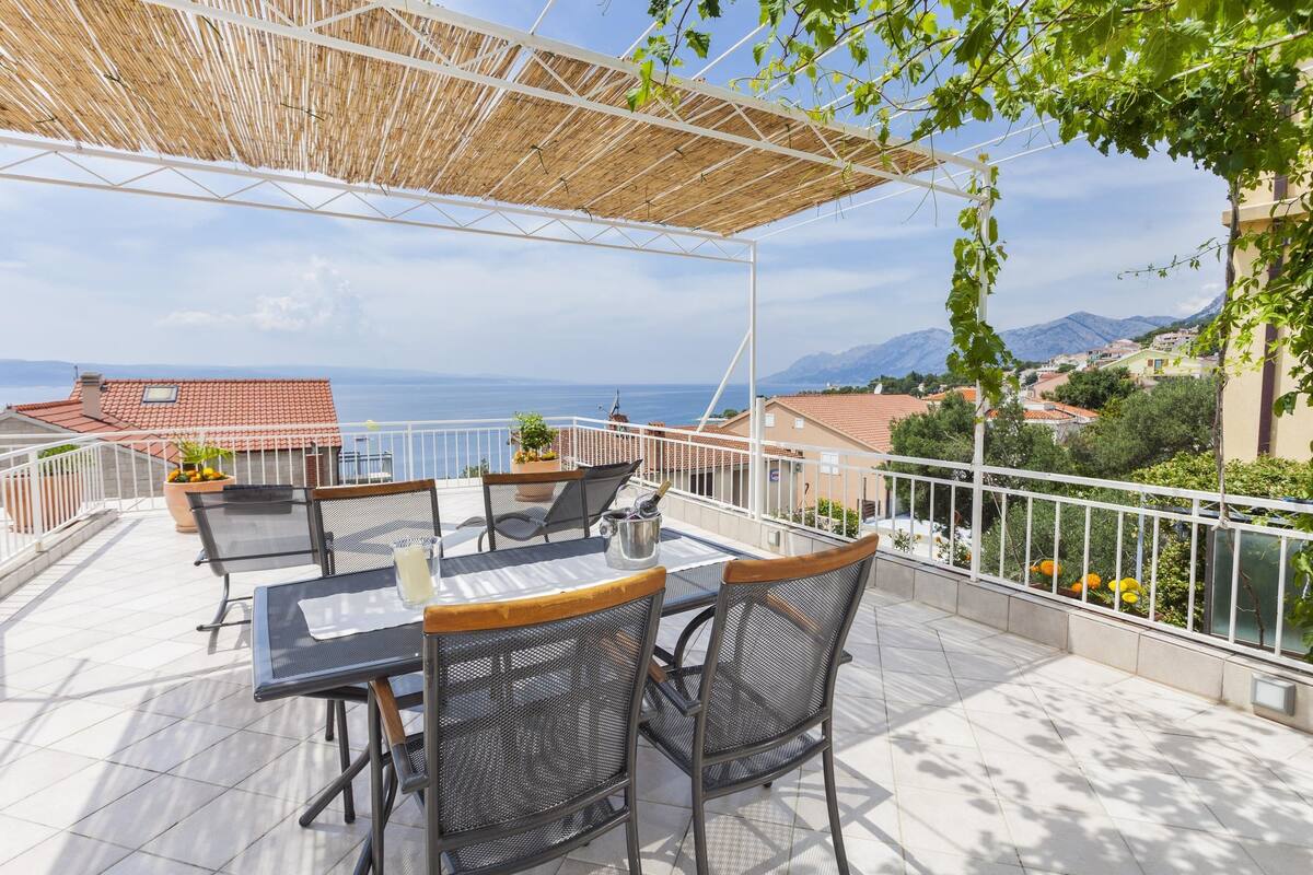 A terrace with a table and chairs, a pergola, and a view of the sea and mountains.