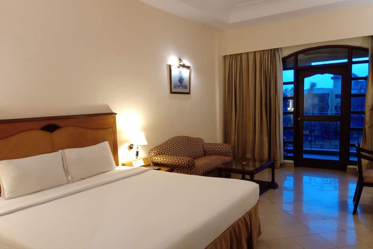 A hotel room with a large bed, a chair, a small table, a lamp, and a view of the outside through a door.