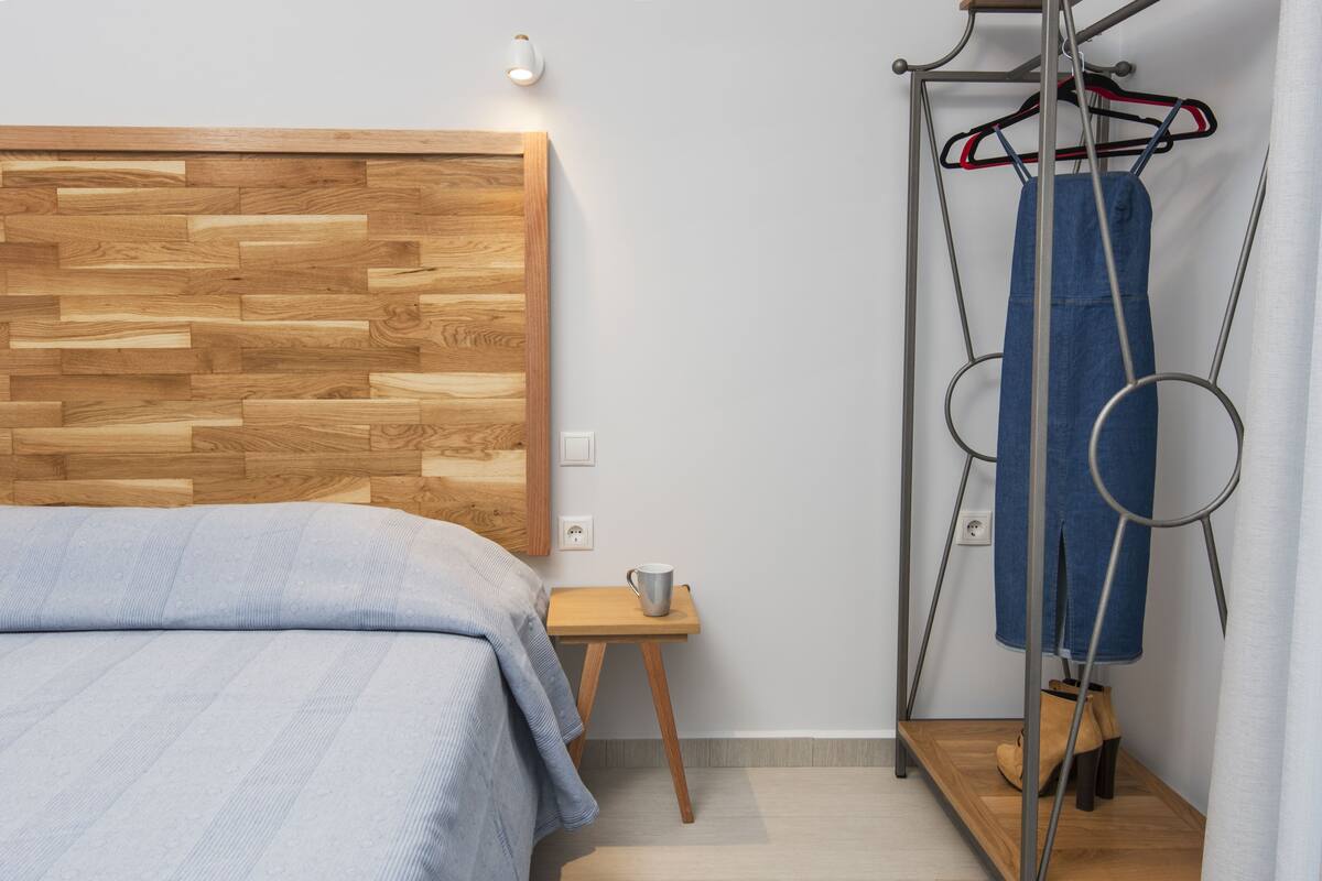 A bedroom with a wooden headboard, a small bedside table with a mug, and a clothing rack with jeans and boots.