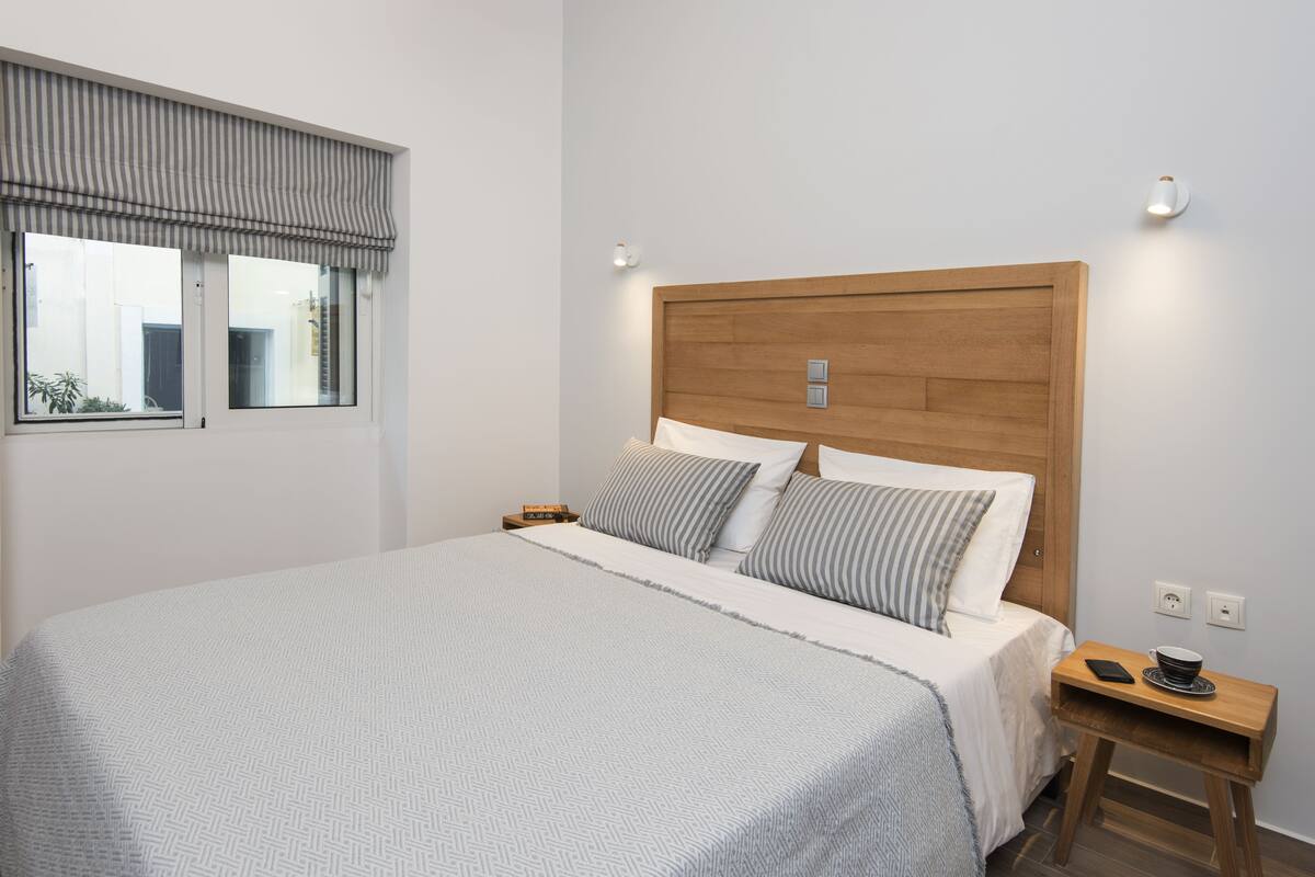 A neatly made bed with a wooden headboard, a bedside table with a phone and a bowl, and a window with a striped blind.