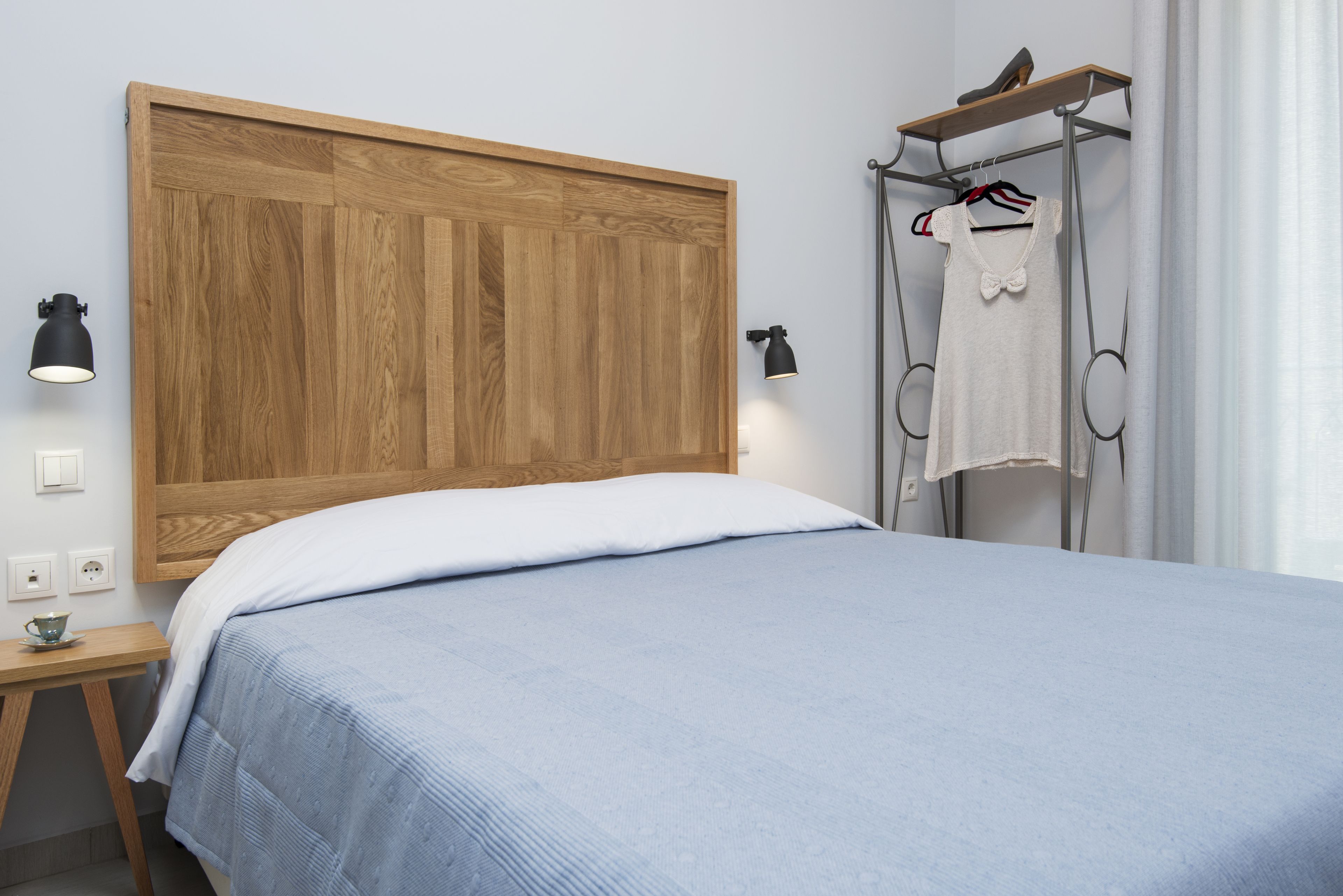Um quarto com cabeceira de madeira, cama com colcha azul, mesa de cabeceira com abajur e um cabideiro com uma camisa branca.