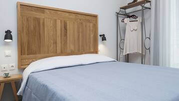 Um quarto com cabeceira de madeira, cama com colcha azul, mesa de cabeceira com abajur e um cabideiro com uma camisa branca.