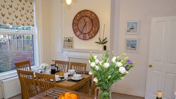 A dining area with a wooden table, chairs, a vase with flowers, a bowl of oranges, and a clock on the wall. - Jutes House Exeter (Exeter)