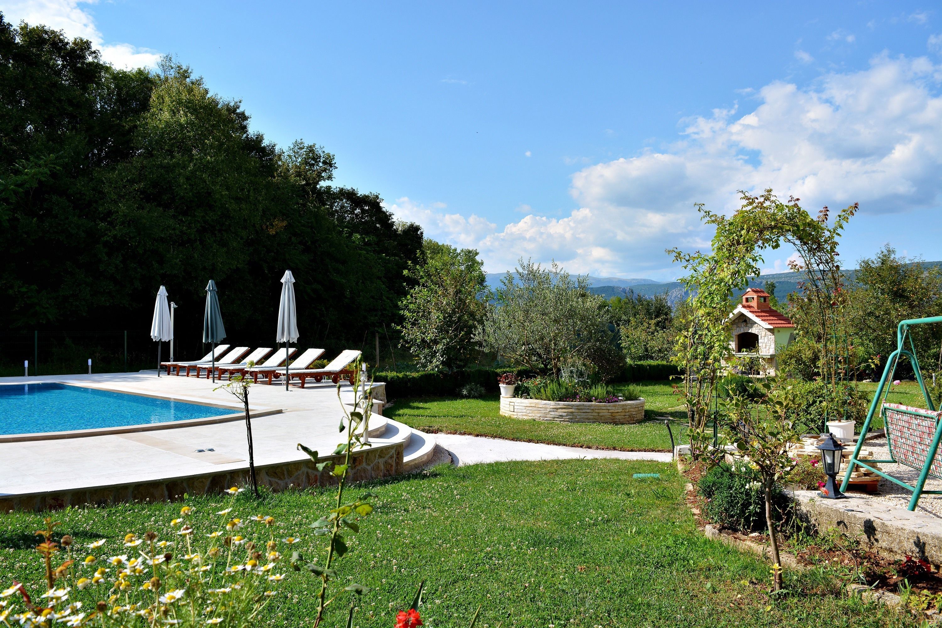 Une piscine entourée de chaises longues et de parasols, elle-même bordée d’un jardin bien entretenu et offrant une vue sur les montagnes.