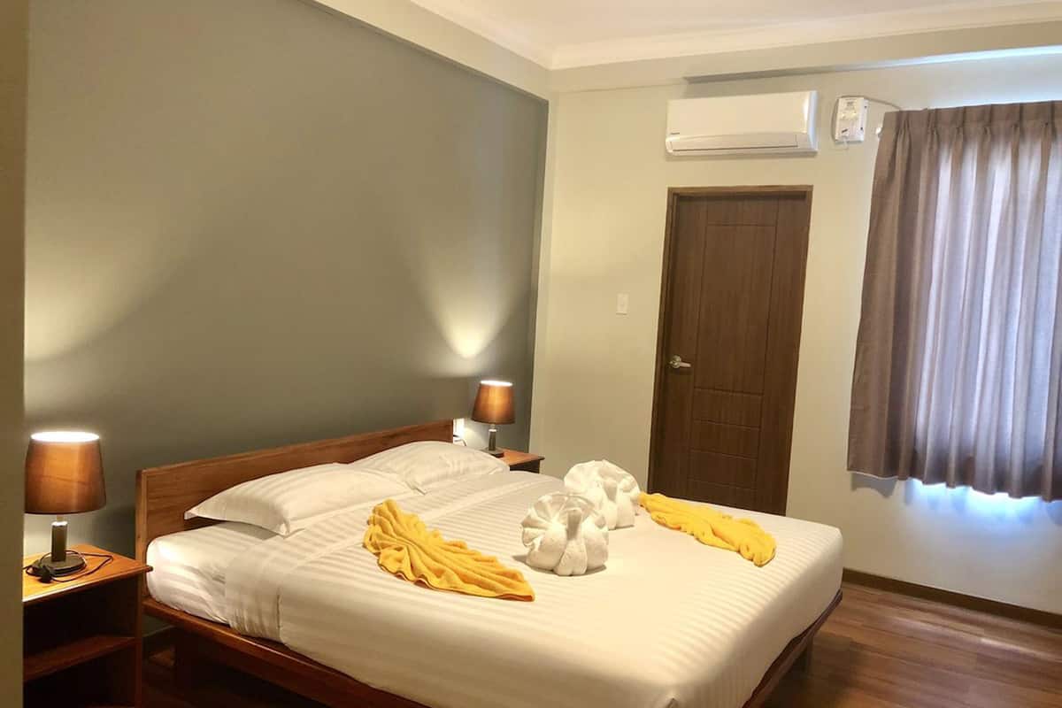 Cama bem arrumada com lençóis brancos, duas luminárias de cabeceira, um ar condicionado e uma janela com cortinas.
