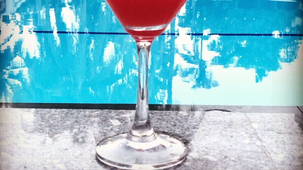 Un cocktail in un bicchiere da martini con un ombrello rosa, appoggiato davanti a una piscina.