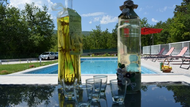 A poolside table with clear glasses and bottles, a pool, lounge chairs, and a red umbrella. - Holiday House 4M (Podbablje)