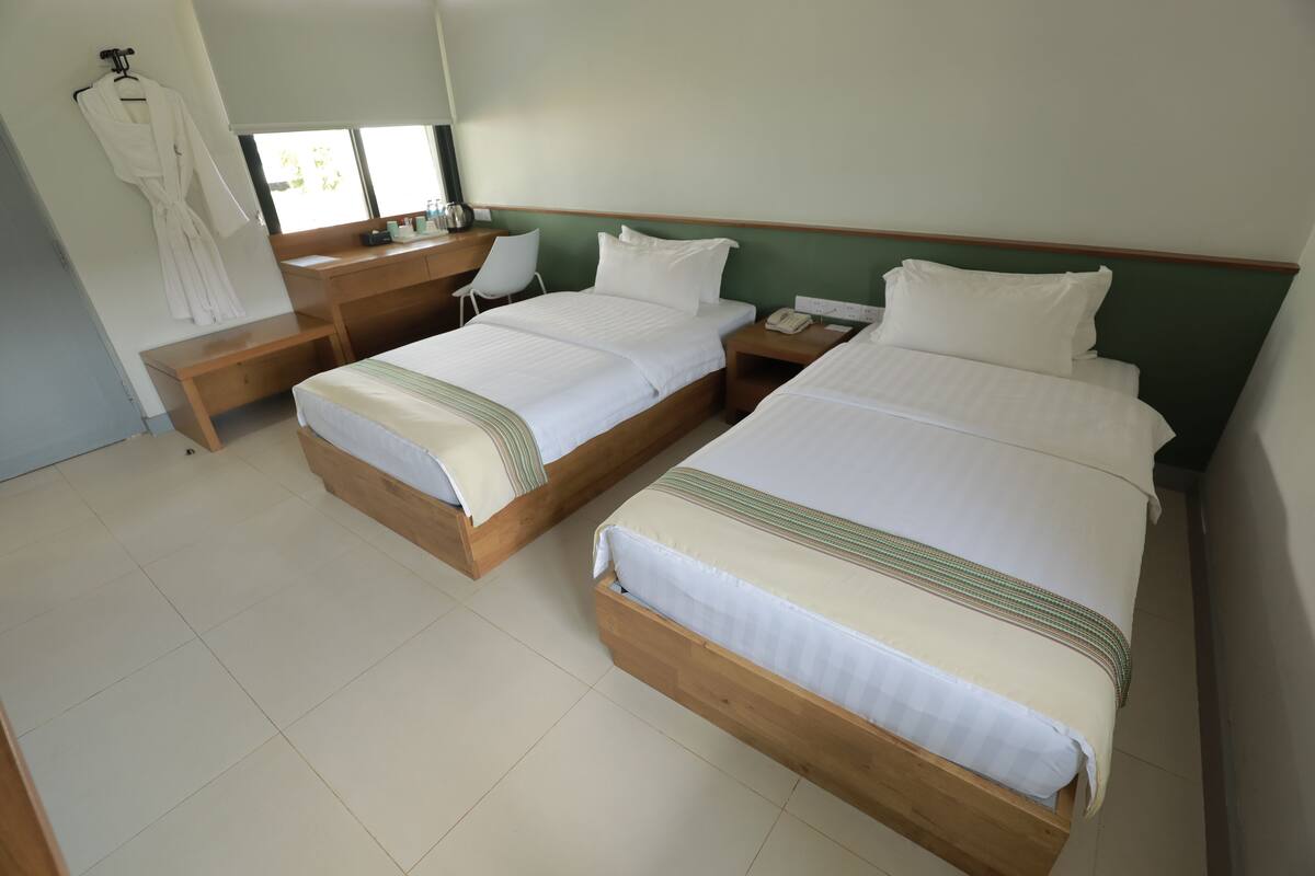 A hotel room with two single beds, a wooden desk, a chair, a robe hanging, and a window with blinds.