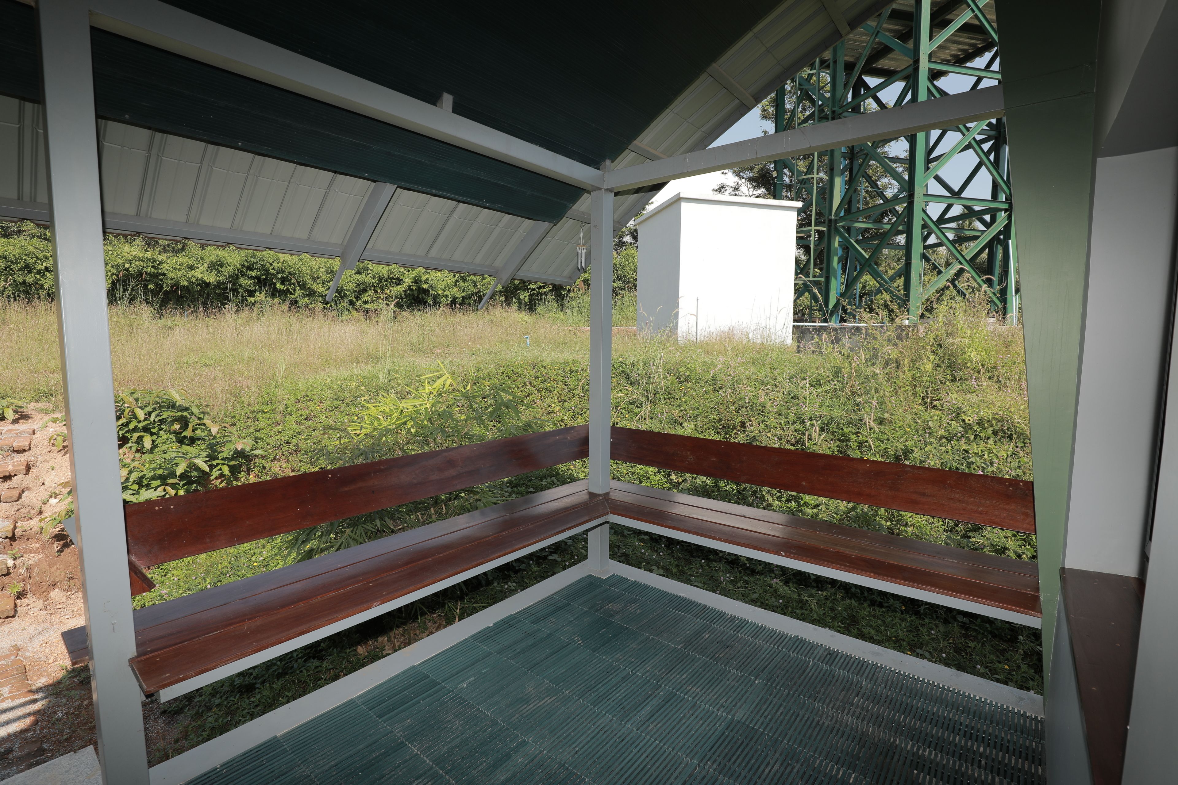 A covered outdoor seating area with wooden benches and a metal floor, overlooking a grassy field and a white building.