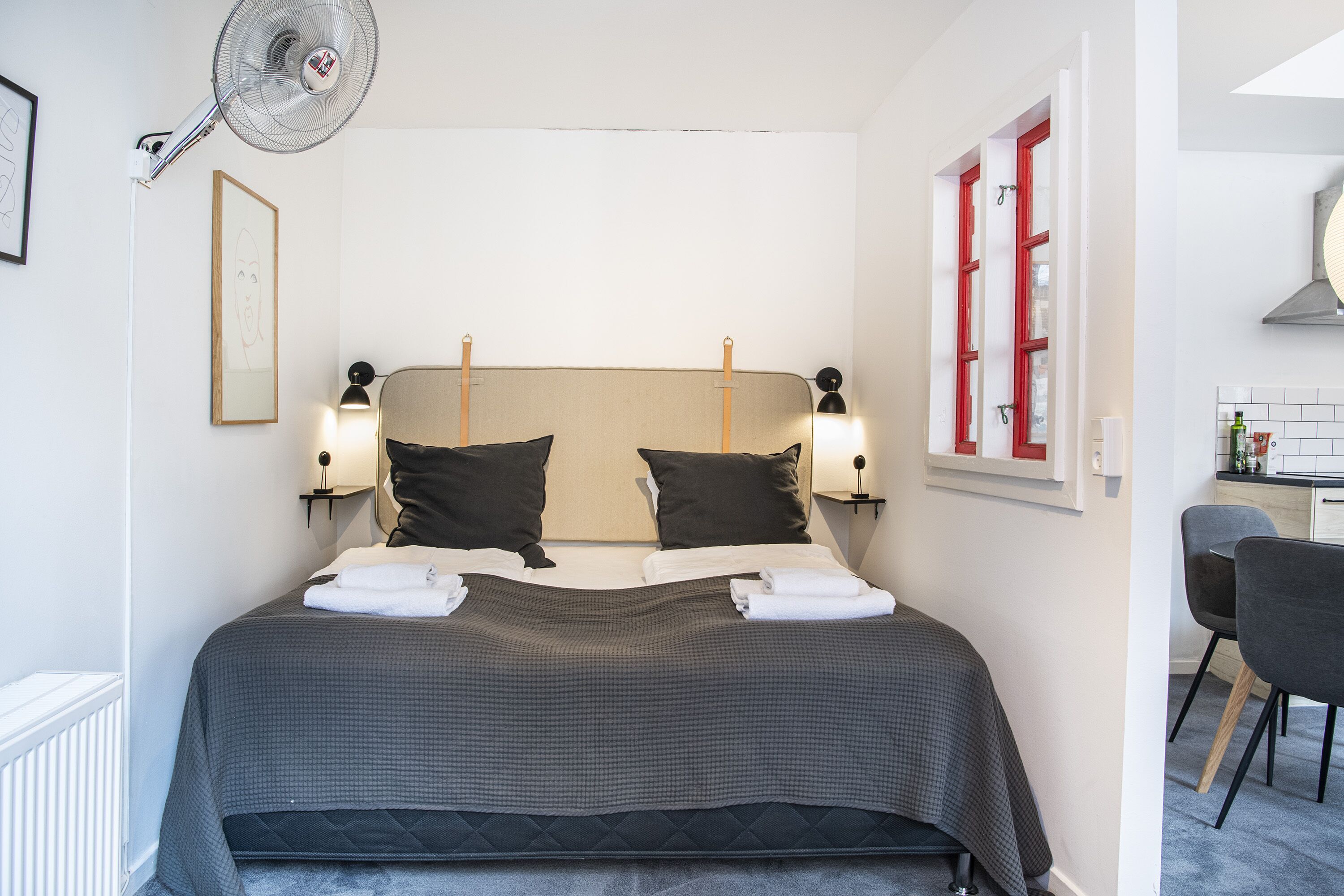 Una cama bien tendida con cabecera, dos lámparas de noche y un ventilador de pared.