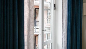 Vista a través de una ventana con marco blanco y picaporte, cortinas entreabiertas y un vistazo a un edificio en el exterior.