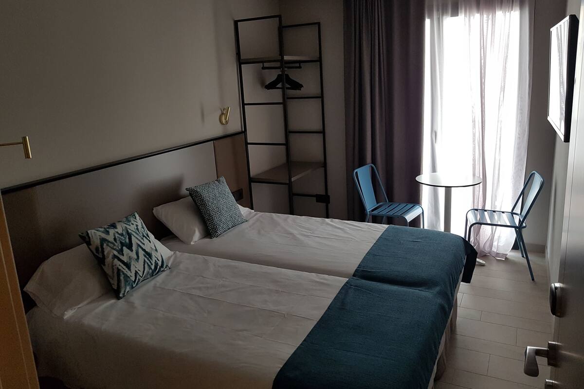 A hotel room with two beds, a small table, two chairs, a wardrobe, and a window with curtains.