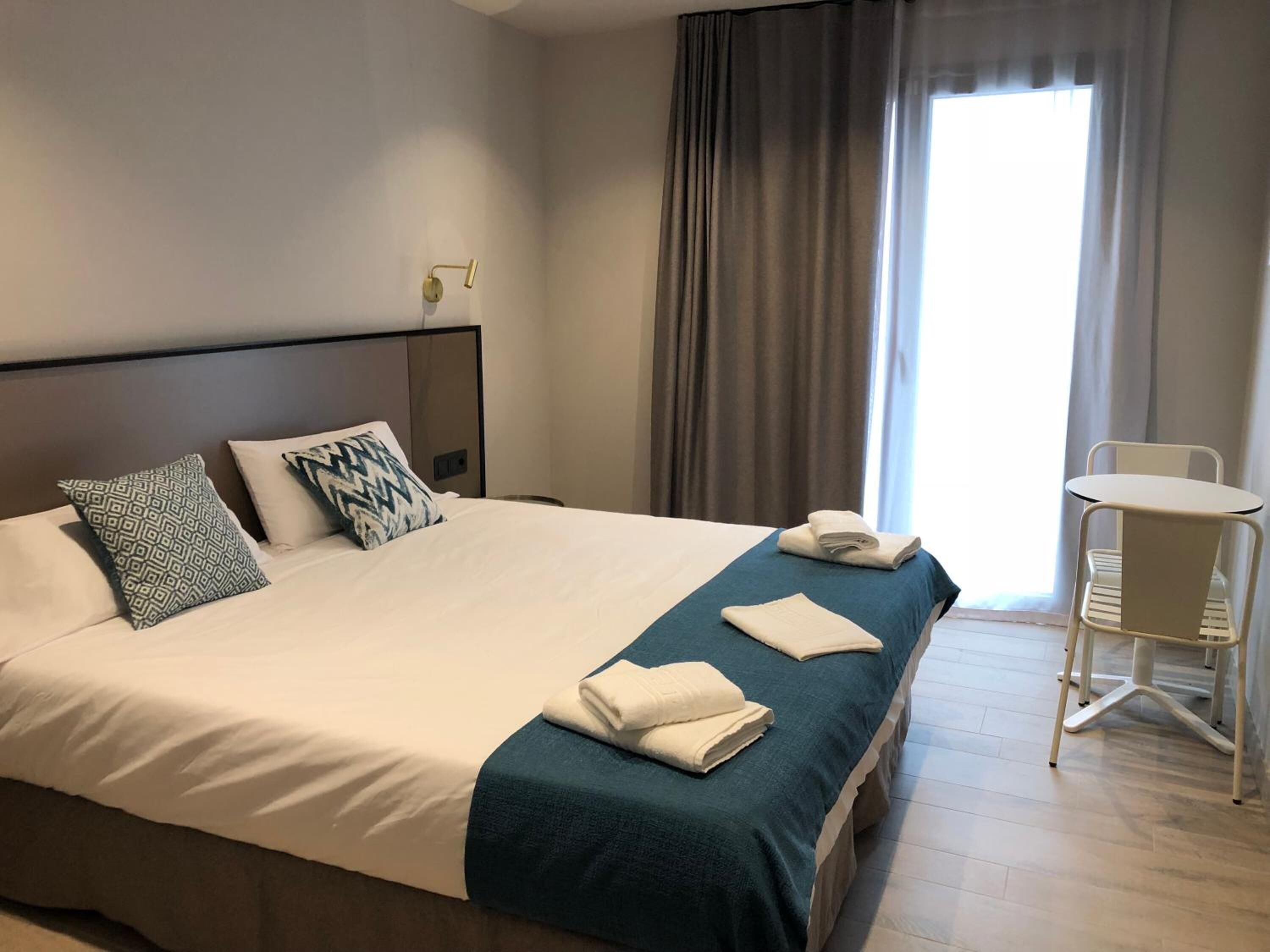 A hotel room with a large bed, two pillows, a bedside table, a chair, and a window with curtains.
