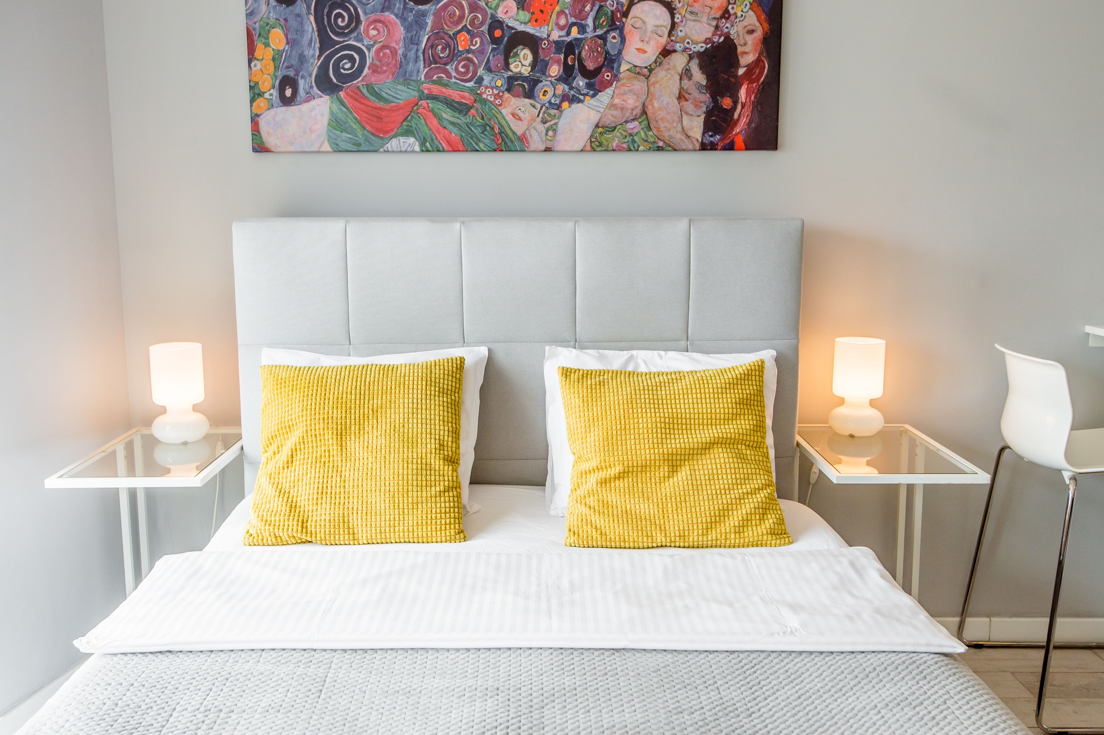 A bed with a grey upholstered headboard, two white pillows, and two yellow pillows. There are two bedside tables with white lamps.