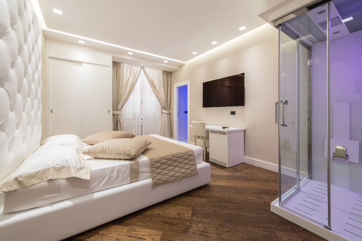 A modern bedroom with a bed, a desk, a TV, and a glass shower enclosure.