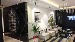 Un moderno vestíbulo de hotel con un mostrador de recepción de mármol negro, una gran araña y una zona de estar con sofás blancos y cojines a rayas.