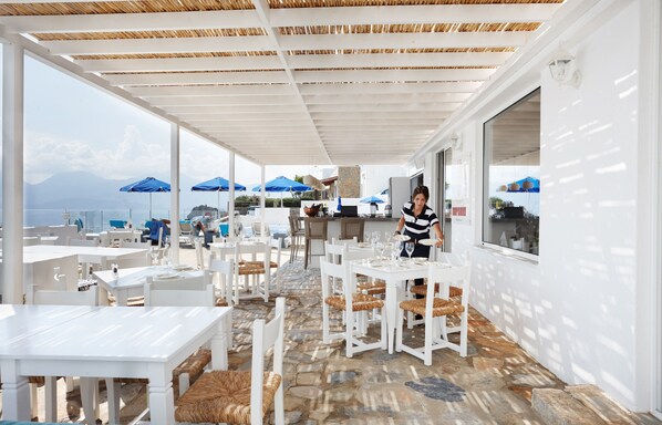 An outdoor dining area with white tables and chairs, a view of the sea, and blue umbrellas. - Karma Minoan (Agios Nikolaos)