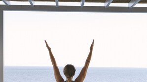 A person performing a yoga pose on a green mat in a room with a view of the ocean. - Karma Minoan (Agios Nikolaos)