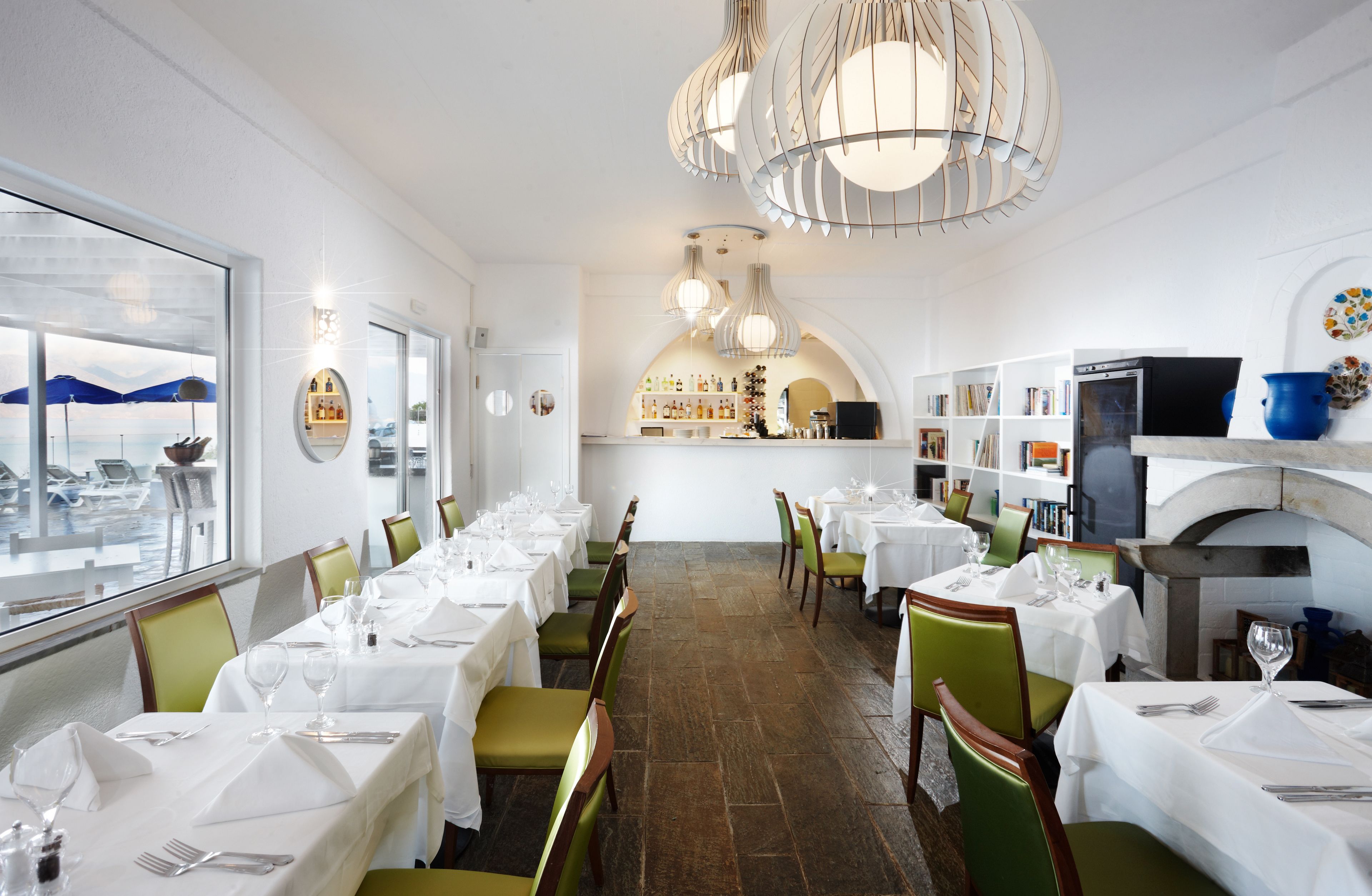 A restaurant with white tables, green chairs, and a view of boats through the window.