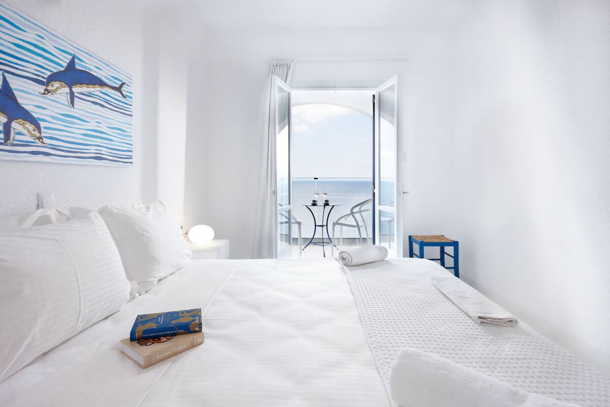 A hotel room with a bed, a small table, a chair, a painting of dolphins, and a view of the sea.