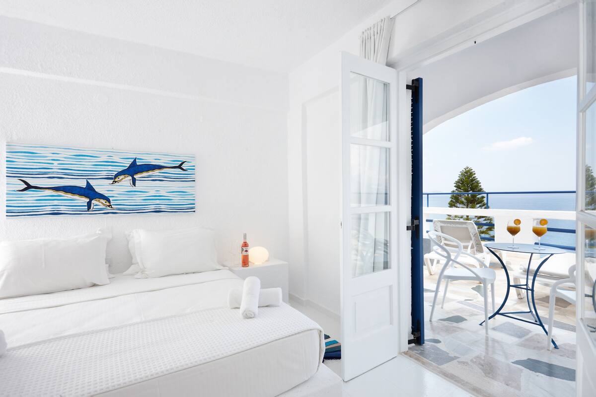 A bright, white room with a bed, a painting of dolphins, and a balcony with a table and chairs.