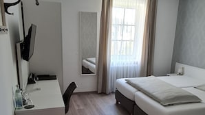 A compact hotel room with a bed, a desk, a chair, a mirror, and a window with curtains. - Hotel Restaurant Franziska (Straubing)
