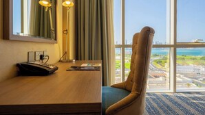 Ein Hotelzimmer mit Schreibtisch, zwei Lampen, einem Sessel und einem Spiegel, mit großem Fenster und Blick auf die Stadt.