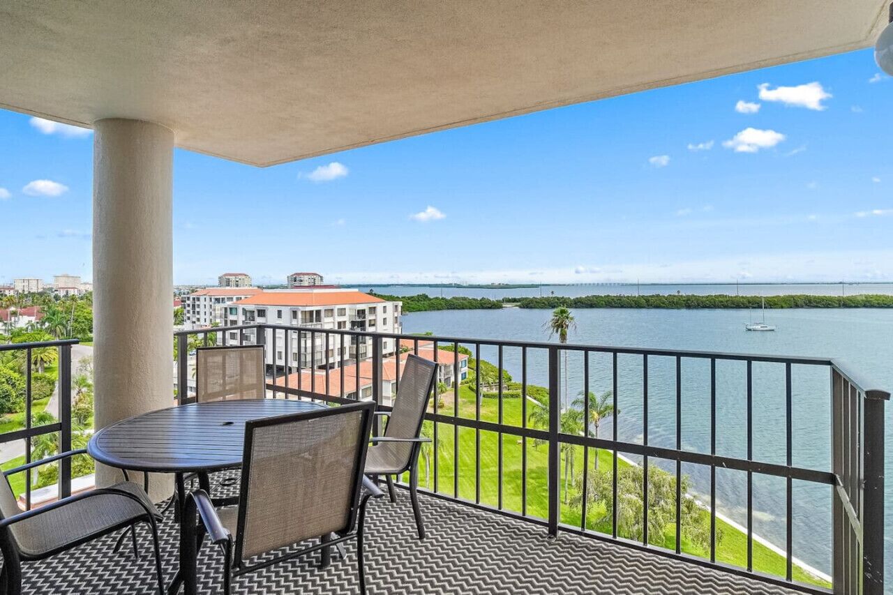En balkon med et bord og stole, ud til et vandområde og en bygning i det fjerne.