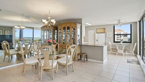 A spacious dining area with a glass-top table surrounded by chairs, a wooden cabinet with glass doors, and a view of the outdoors through large windows. - Penthouse Paradise with Amazing Views: 2BD, 2BA - (Isla Del Sol)