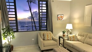 A living room with a large window, a sofa, an ottoman, a side table with a lamp, and a potted plant. - Spectacular  Ocean Front 1 Bedroom Villa in Golf community, Pal mas Del Mar  (Palmas Del Mar)