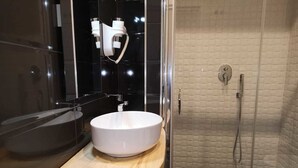 A modern bathroom with a white sink, a wooden vanity, a glass shower enclosure, and a black tiled shower wall. - Beverello Rooms (Naples)