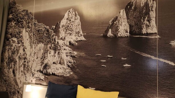 A room with a large wall mural of a coastal landscape, a bed with pillows, and a bedside table with a lamp. - Beverello Rooms (Naples)