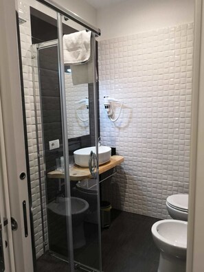 A modern bathroom with a glass shower enclosure, a white tiled wall, a sink with a wooden countertop, and a toilet. - Beverello Rooms (Naples)