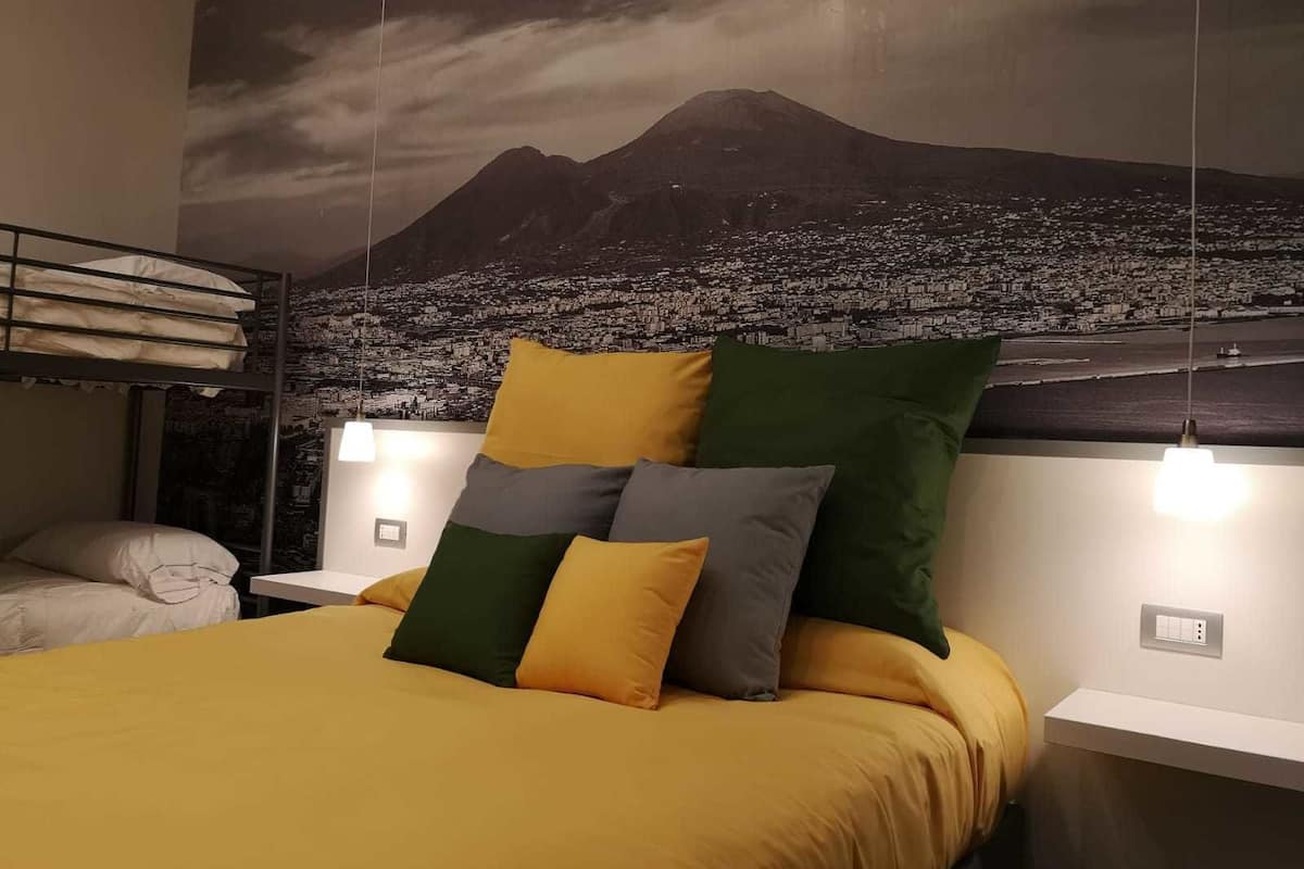 A bed with a yellow bedspread and pillows, a bunk bed to the left, and a cityscape mural on the wall.