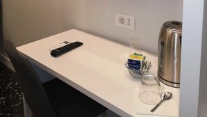 A small white desk with a TV mounted above it, a remote control, a kettle, and some toiletries on the counter. - Beverello Rooms (Naples)