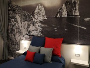 A bedroom with a bed, pillows, a nightstand, and a wall mural of a coastal scene. - Beverello Rooms (Naples)